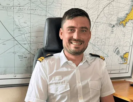 Uniformed officer seated at a desk in an office, with a large maritime navigation chart mounted on the wall behind.