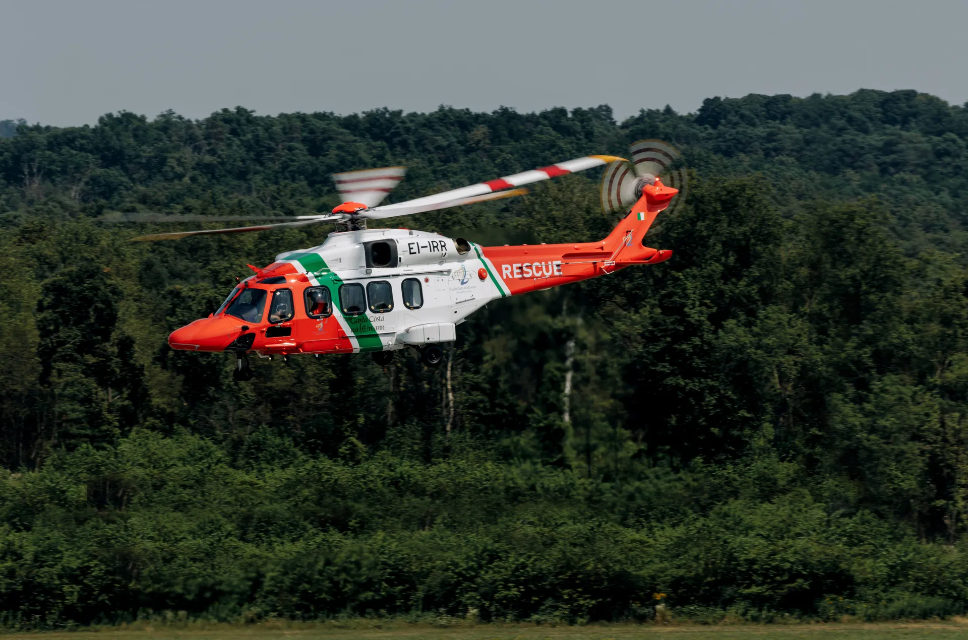 Rescue helicopter flying at low altitude over land, with forested terrain in the background.