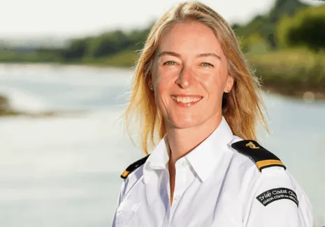 Uniformed officer standing outdoors near calm water, with shoreline and greenery visible in the background.