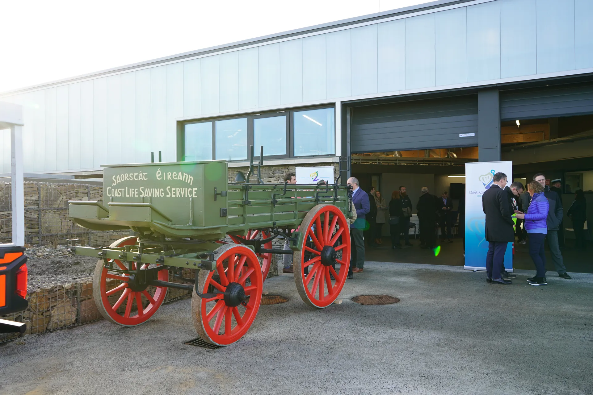 Historic horse-drawn life-saving cart displayed outside a modern coastal facility, with people gathered nearby.