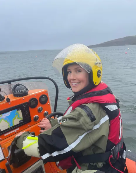 Crew member wearing a helmet and flotation gear seated at the controls of a small rescue boat, with open water and coastline visible behind.