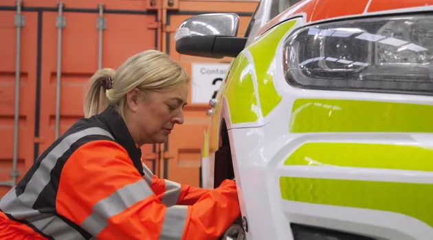 A rescue service worker in high-visibility clothing carries out maintenance on a marked emergency response vehicle inside a depot or garage.
