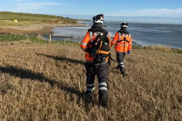 Two personnel in high visibility jackets walking across coastal grassland towards the shoreline under clear skies.