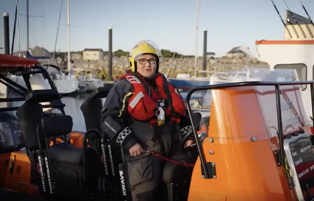 Crew member wearing a helmet and flotation gear standing aboard a small rescue boat moored in a marina, with other vessels visible in the background.