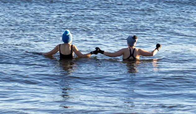 Two people holding hands while standing chest deep in the sea, wearing swimwear and hats in cold water conditions.
