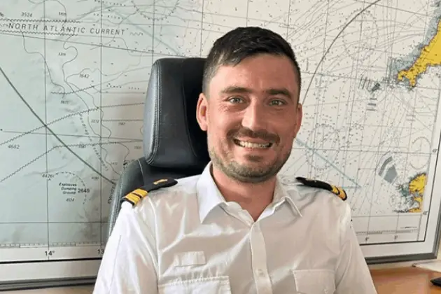 Uniformed officer seated at a desk in an office, with a large maritime navigation chart mounted on the wall behind.