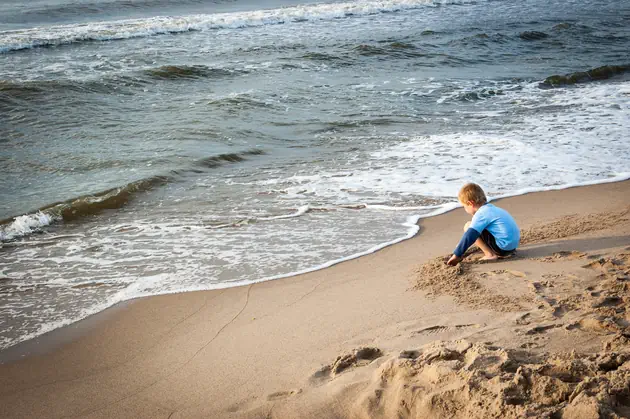 A child sits on a sandy beach near the water’s edge, playing in the sand as gentle waves roll onto the shore.