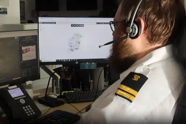 Uniformed officer seated at a workstation, viewing maritime information on dual computer screens in a control room setting.