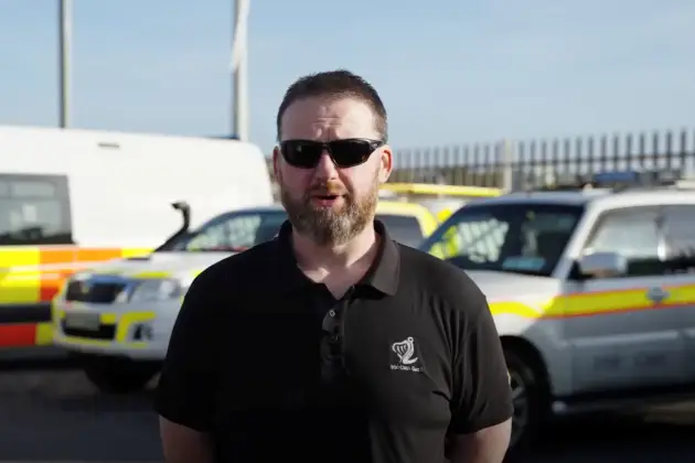 Person standing outdoors in front of emergency response vehicles parked behind a secure fence.