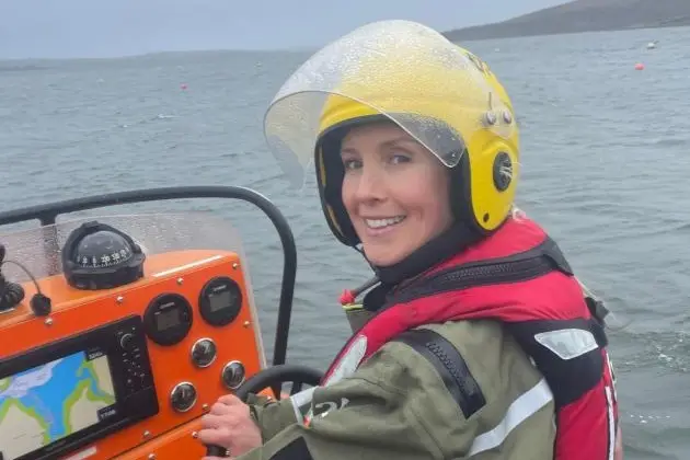 Crew member wearing a helmet and flotation gear seated at the controls of a small rescue boat, with open water and coastline visible behind.