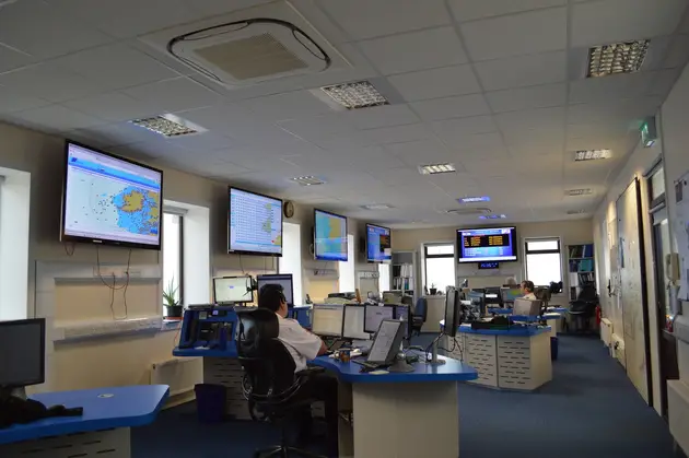 Operations room with personnel seated at workstations, monitoring maritime charts and information displayed on wall mounted screens.