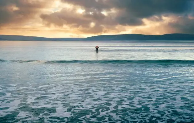 Single swimmer standing in calm open water at a distance from shore, with low waves and clouds overhead.