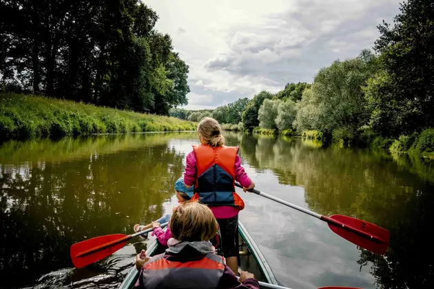 Two children wearing flotation vests paddling a canoe along a calm river bordered by trees.