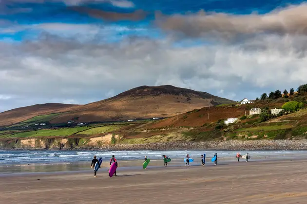 People carrying surfboards walking along a wide sandy beach, with rolling waves and hills rising in the background.