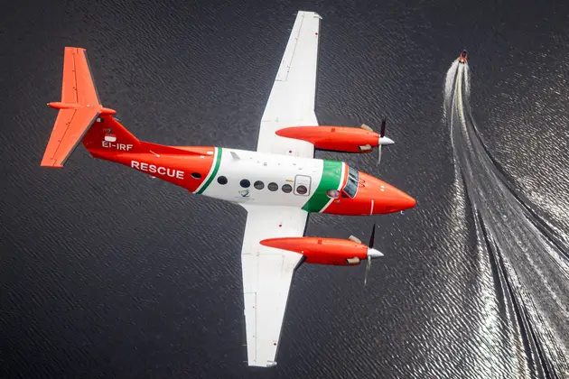 Aerial view of a twin‑engine rescue aircraft flying over open water, with a small vessel below.