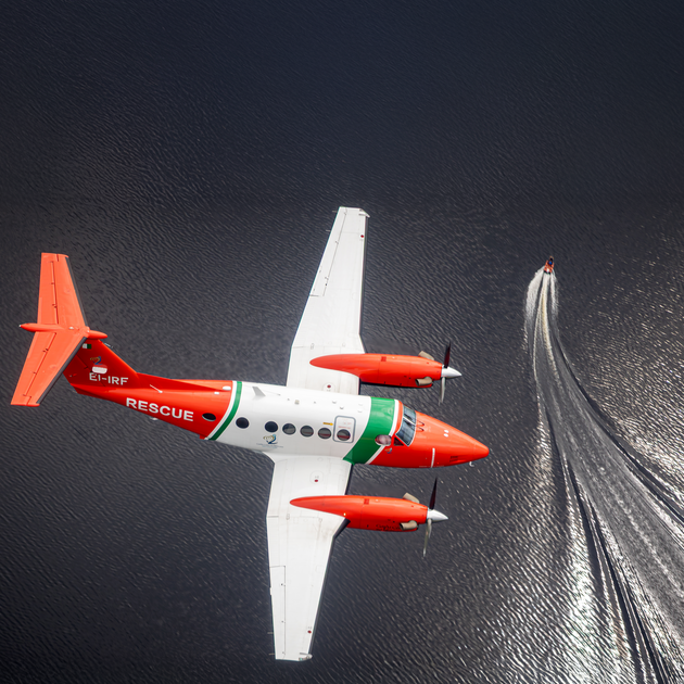 A red, white and green rescue aircraft flies over dark water, with a small speedboat nearby leaving a sweeping white wake across the surface
