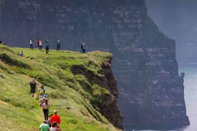 People walking along a coastal cliff path above the sea, with steep rock faces and ocean below.