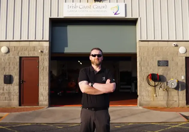 Person standing with arms crossed outside a coastal operations building, with an open vehicle bay visible behind.