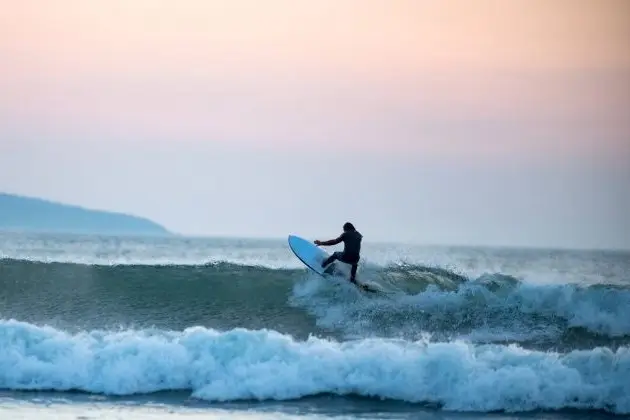 Surfer riding a breaking wave offshore, with coastline visible in the background.