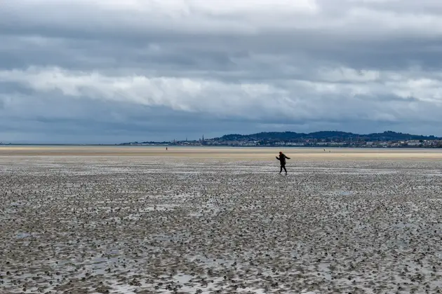 Wide tidal flat at low tide with a lone person walking across wet sand, distant shoreline visible under heavy clouds.