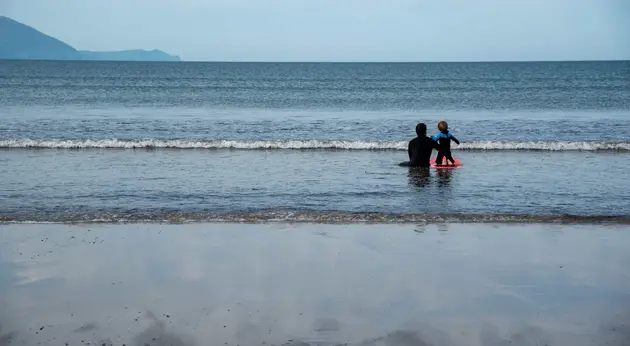 Adult and child seated together in shallow water at the shoreline, facing the open sea.