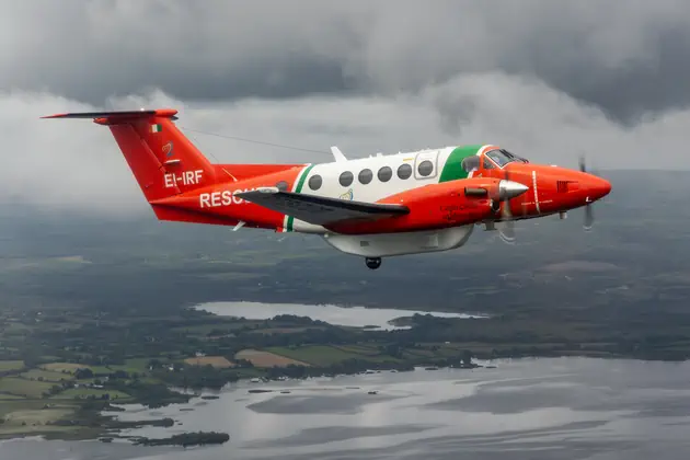 A red, white and green rescue aircraft flies above the Irish countryside under dark clouds, with patchwork fields visible below.