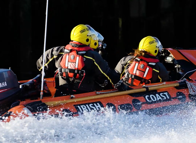 Two Irish Coast Guard crew members in yellow helmets and lifejackets travel in a rescue boat, with sea spray rising around the vessel