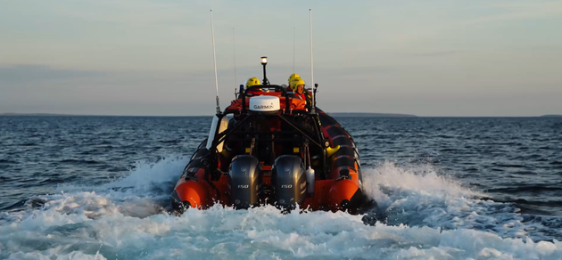 A rescue boat with crew in safety gear speeds across the sea, leaving a trail of wake behind it.